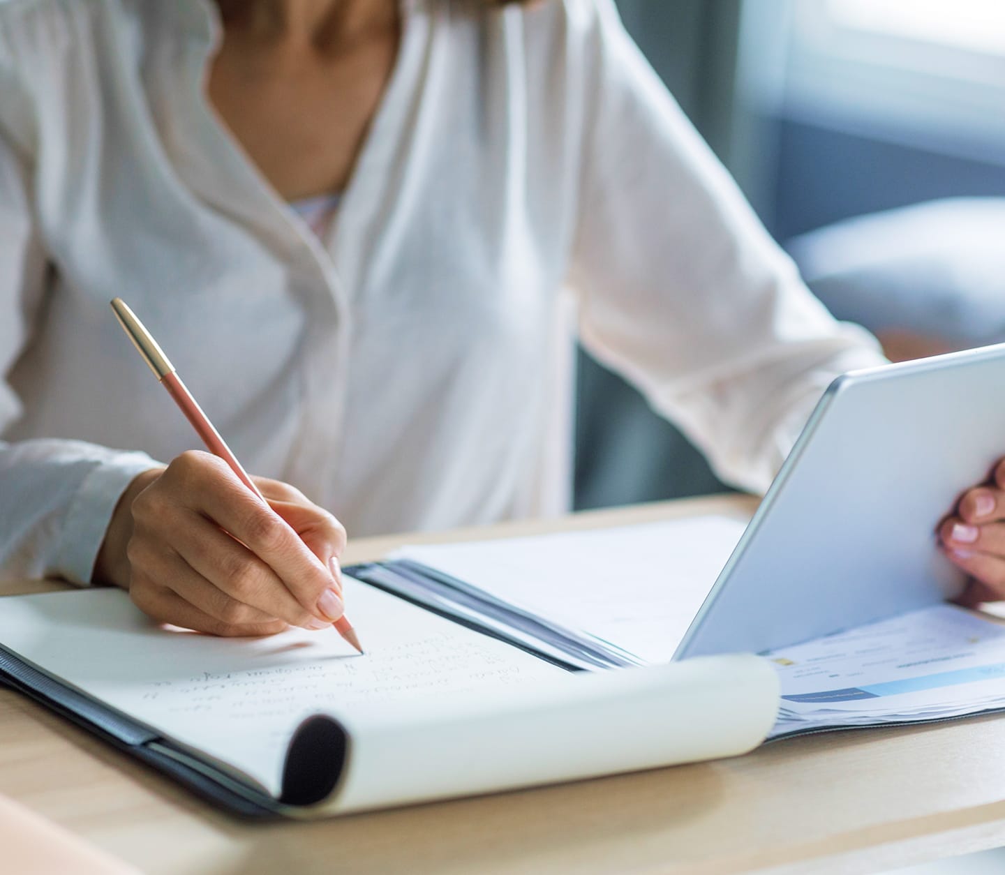 Person writing notes with a pencil in a notebook while holding a tablet.
