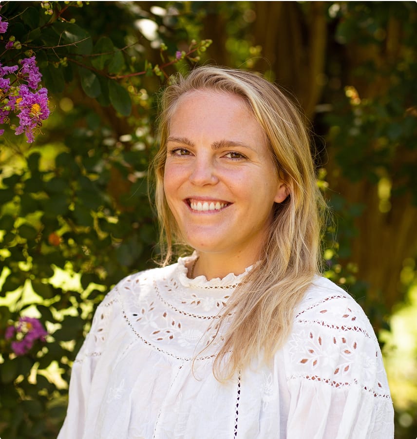 Smiling woman with long blonde hair wearing a white blouse standing outdoors near green foliage and purple flowers.