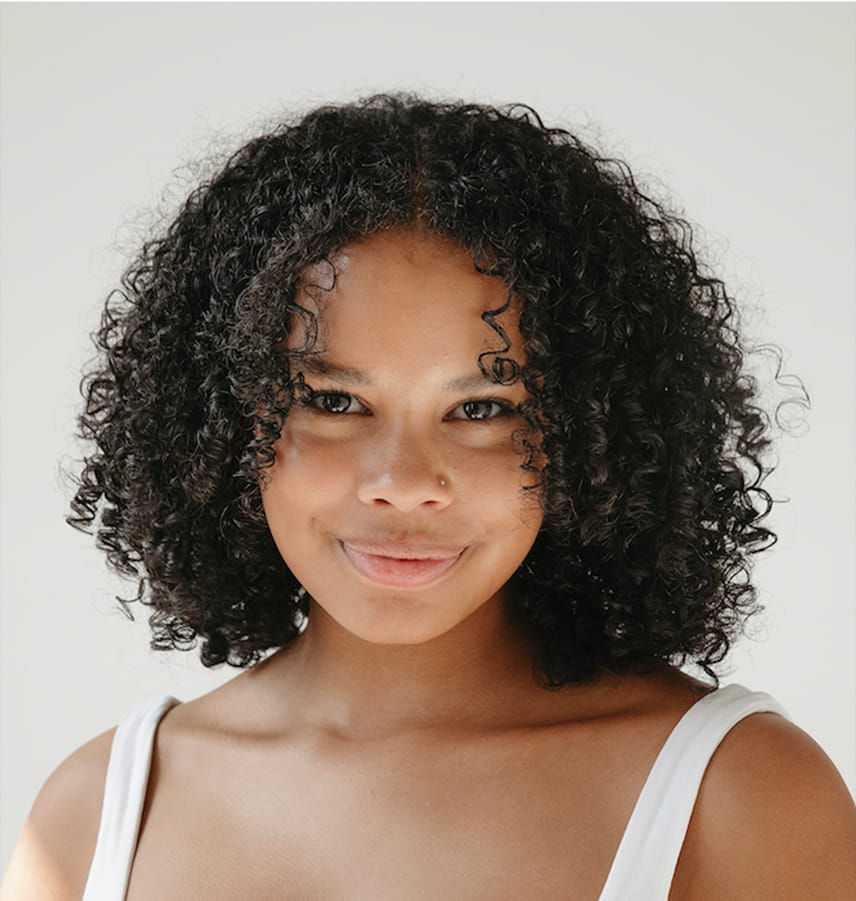 Smiling young woman with curly dark hair wearing a white tank top against a light background.
