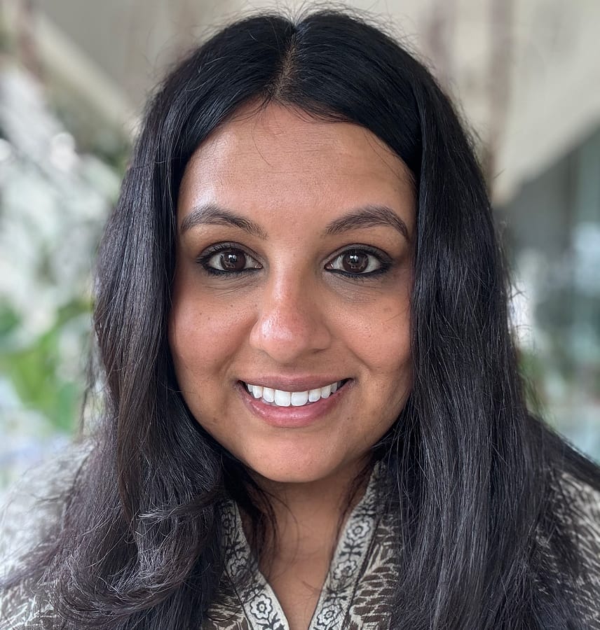 Smiling woman with long dark hair wearing a patterned top in a bright indoor setting.