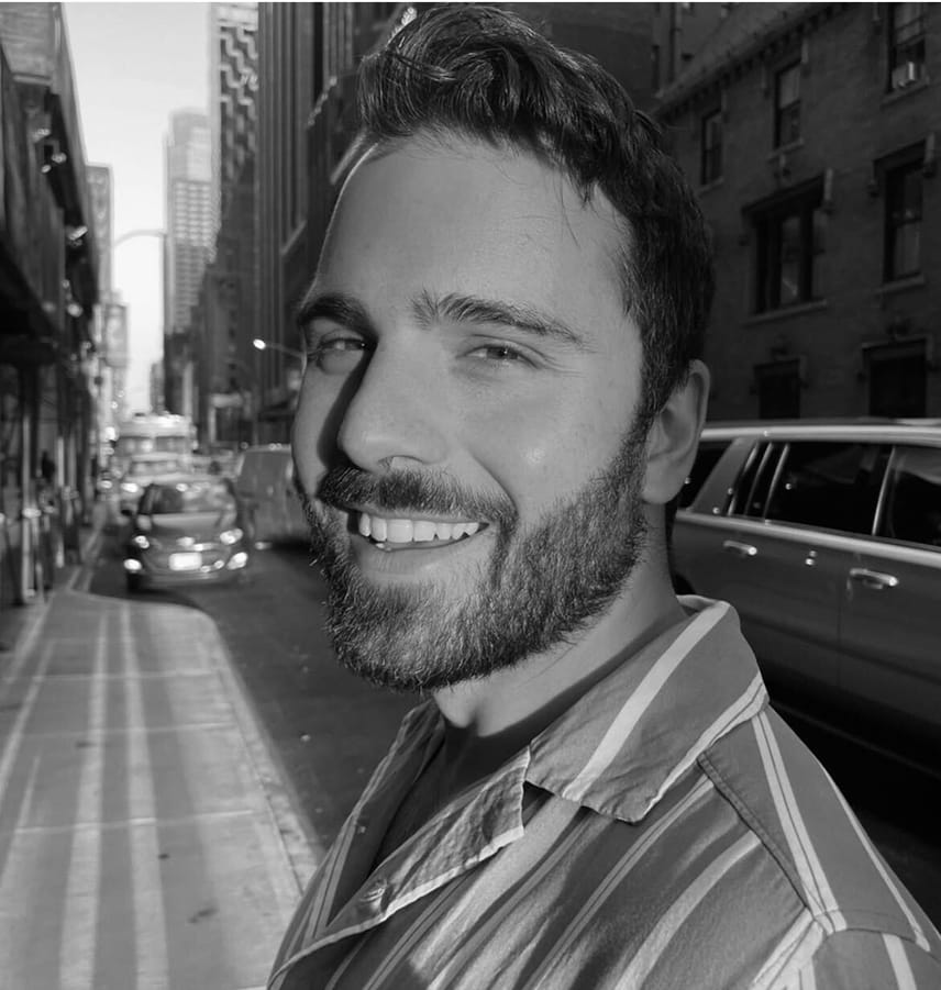 Smiling bearded man wearing a striped shirt standing on a city street with cars and tall buildings in the background.