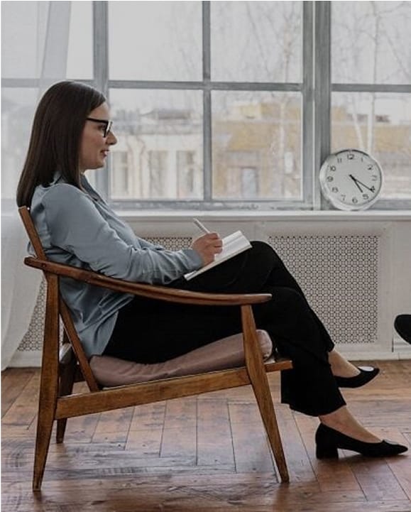 Woman wearing glasses sitting in a wooden chair by a window, writing in a notebook with a clock showing 4:10 in the background.