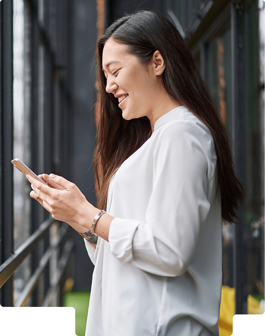 Smiling woman with long black hair wearing white blouse looking at her smartphone indoors by large windows.