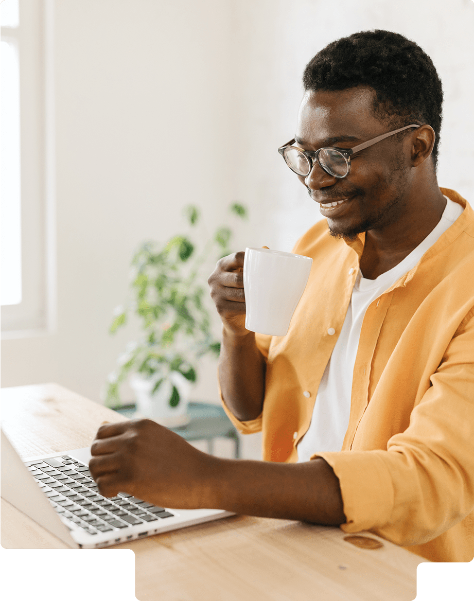 Smiling man wearing glasses and an orange shirt, working on a laptop while holding a white mug.