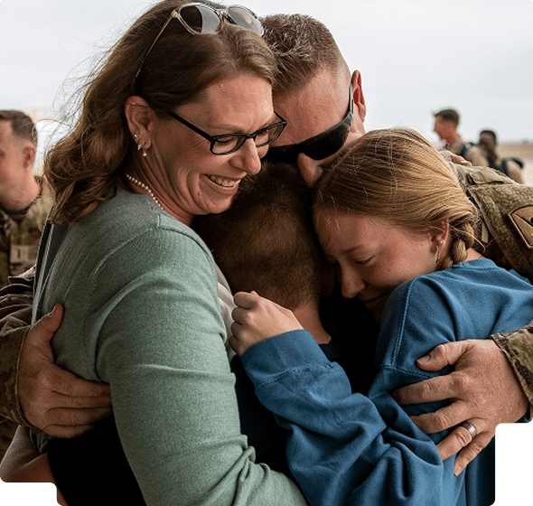 A smiling woman, a man in military uniform, and two children sharing a close group hug outdoors.