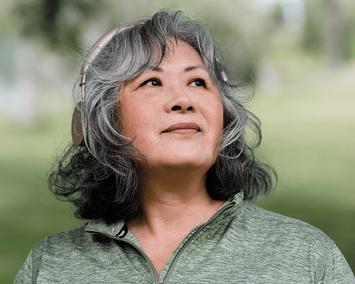 Middle-aged woman with gray hair wearing headphones and a green zip-up shirt looking thoughtfully upward outdoors.