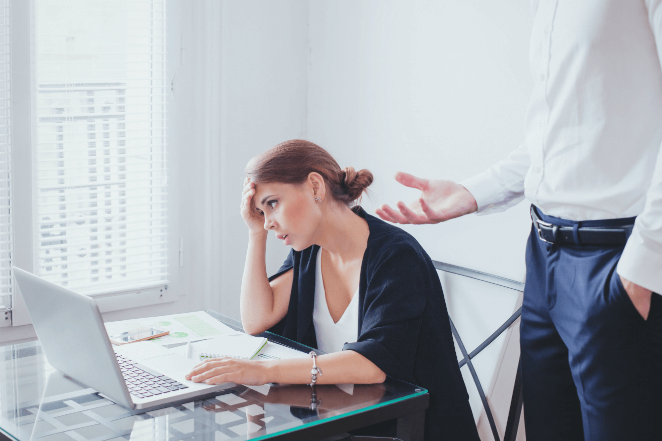 female employee at desk frustrated with male boss standing behind her